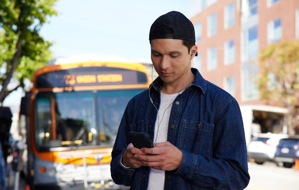 Man wearing earbuds looking down and typing on his phone with a city bus in the background